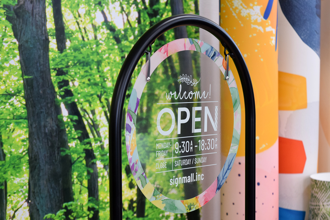完成した透明アクリルのwelcome! OPEN面板のアップ。背景の緑が透けて、水彩フラワーの縁取りとデザインが際立つ
